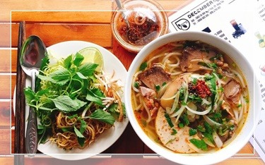 Dwie miski z zupą azjatycką: jedna z makaronem, mięsem i plastrami wędliny, druga z ziołami, kiełkami i makaronem, obok pałeczki, łyżka i słoiczek z przyprawami, w tle menu restauracji.