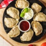 Czarny talerz z ułożonymi pierogami, dipem śmietanowym z bazylią i sosem czerwonym, na drewnianej desce z fragmentem czerwono-białej kraty w tle.