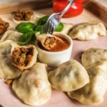 Ręcznie robione pierogi z mięsnym farszem, podane na różowym talerzu z sosem w małej miseczce, udekorowane listkami bazylii i czerwoną papryką w tle.