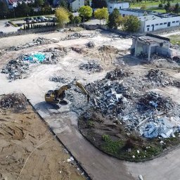 AL-PA sp z o.o. - Widok z lotu ptaka na teren po rozbiórce budynku z gruzem, pracującą koparką i fragmentem ocalałej konstrukcji. W tle zabudowa miejska i parking.