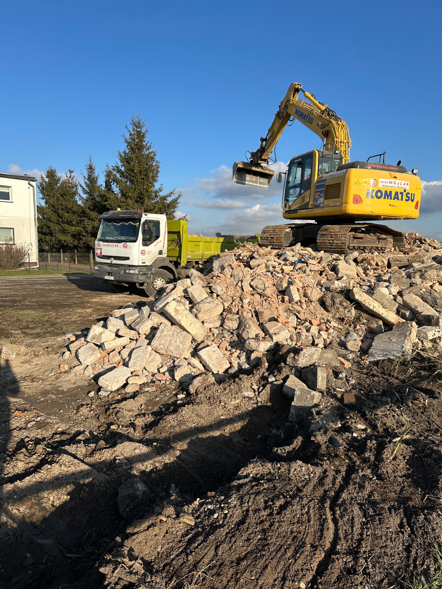 Żółta koparka Komatsu na szczycie gruzowiska z fragmentami cegieł i betonu, obok biała ciężarówka z zieloną skrzynią ładunkową, błękitne niebo w tle.