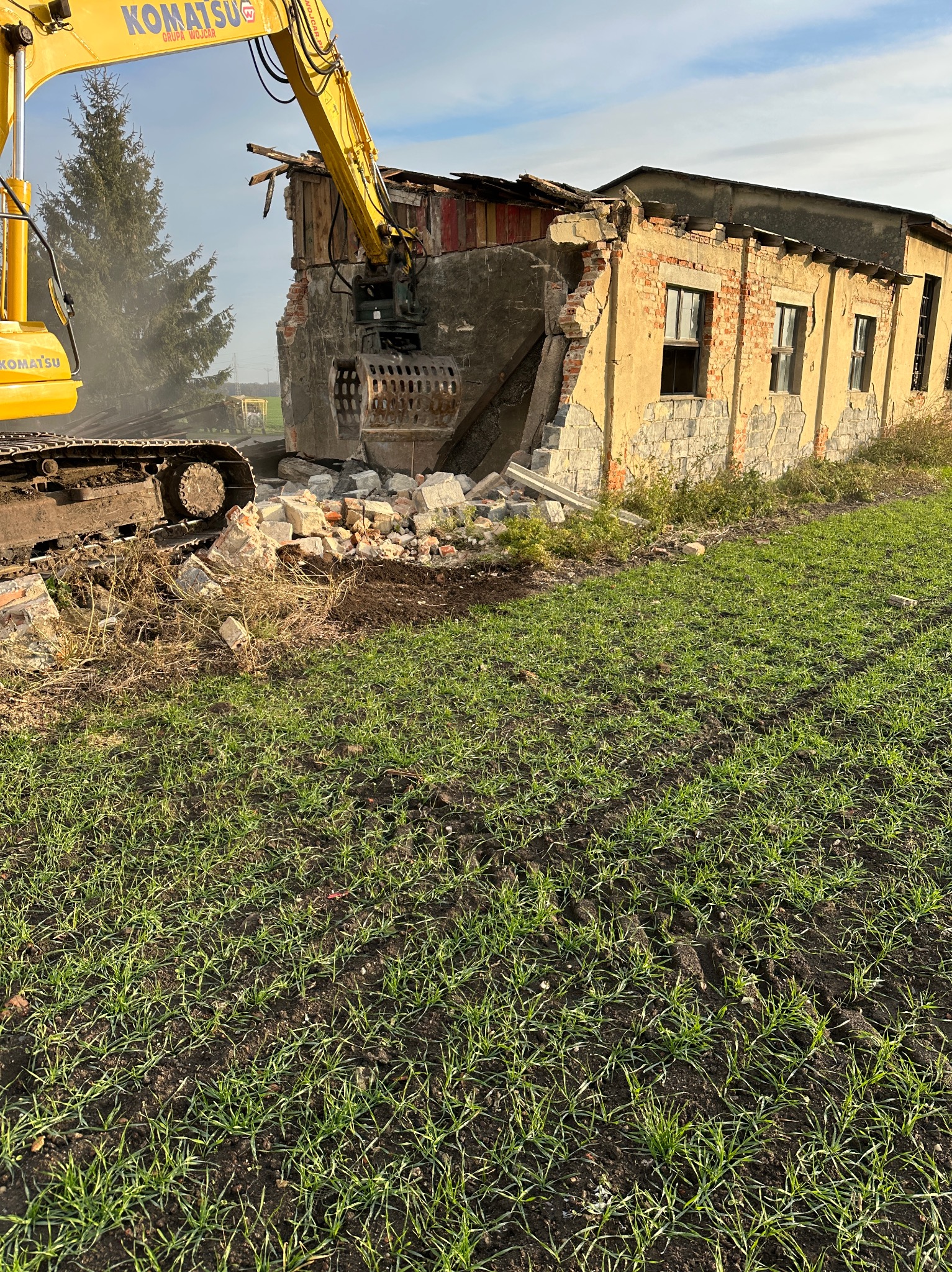 Żółta koparka Komatsu z chwytakiem rozbiera ceglany budynek, fragmenty muru leżą na ziemi, w tle widać drzewo iglaste i błękitne niebo.
