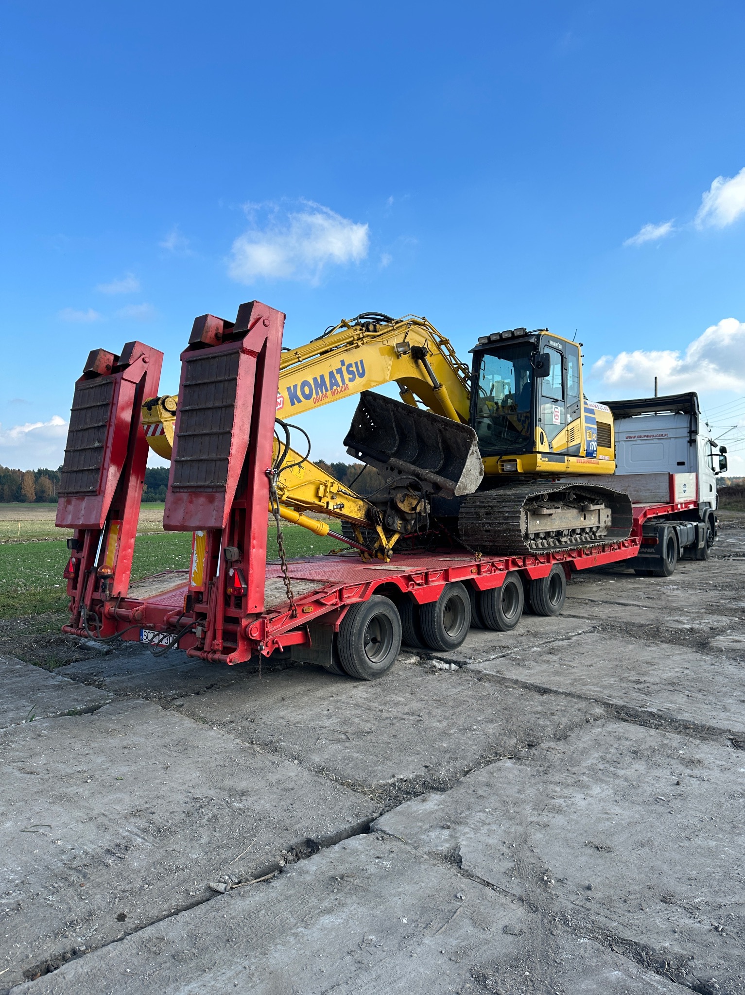 Żółta koparka Komatsu na czerwonej niskopodwoziowej platformie transportowej, widok z tyłu, błękitne niebo z chmurami w tle.