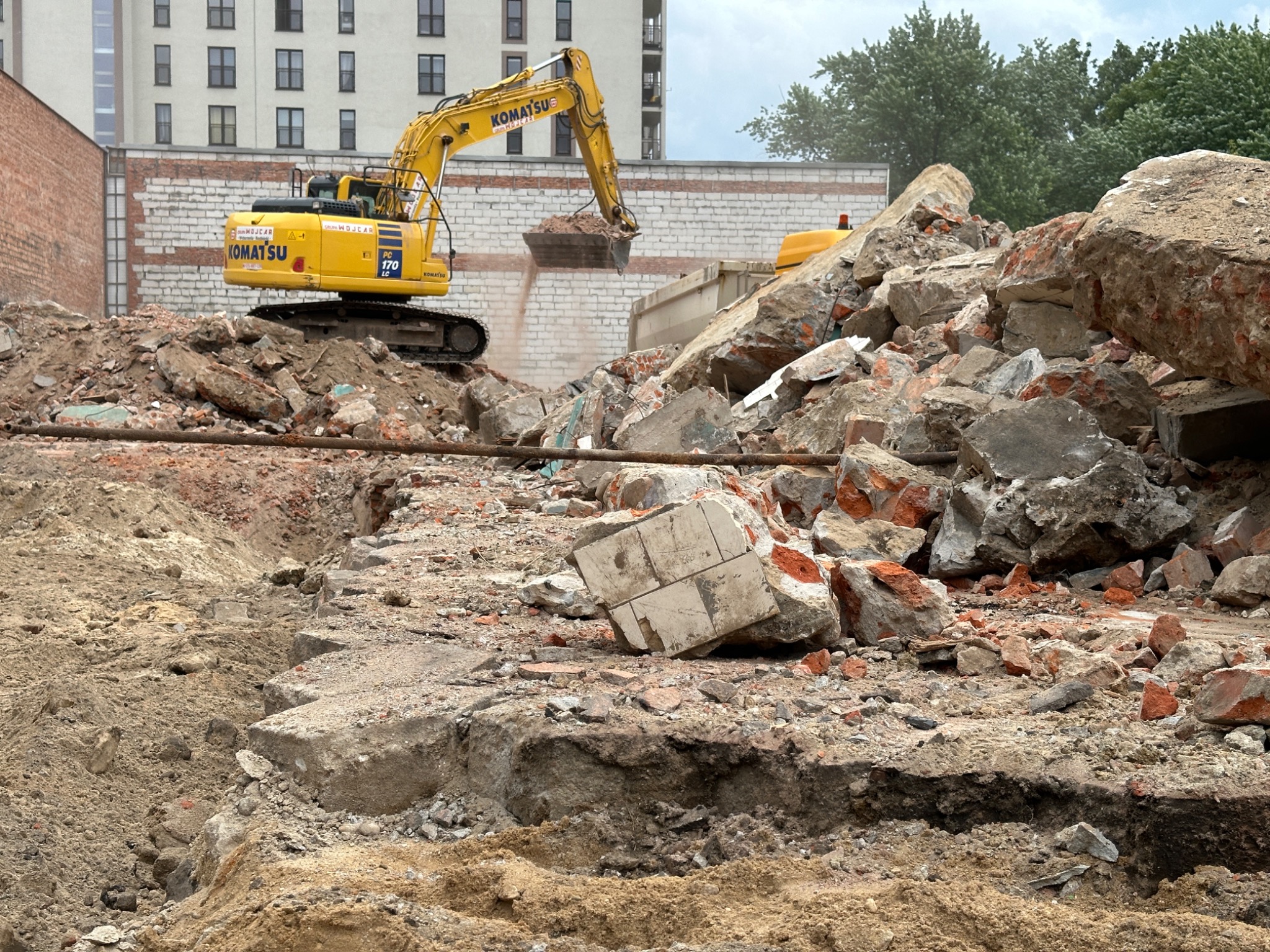 Żółta koparka Komatsu na placu budowy, gruzowisko z cegieł i betonu, w tle budynek mieszkalny.
