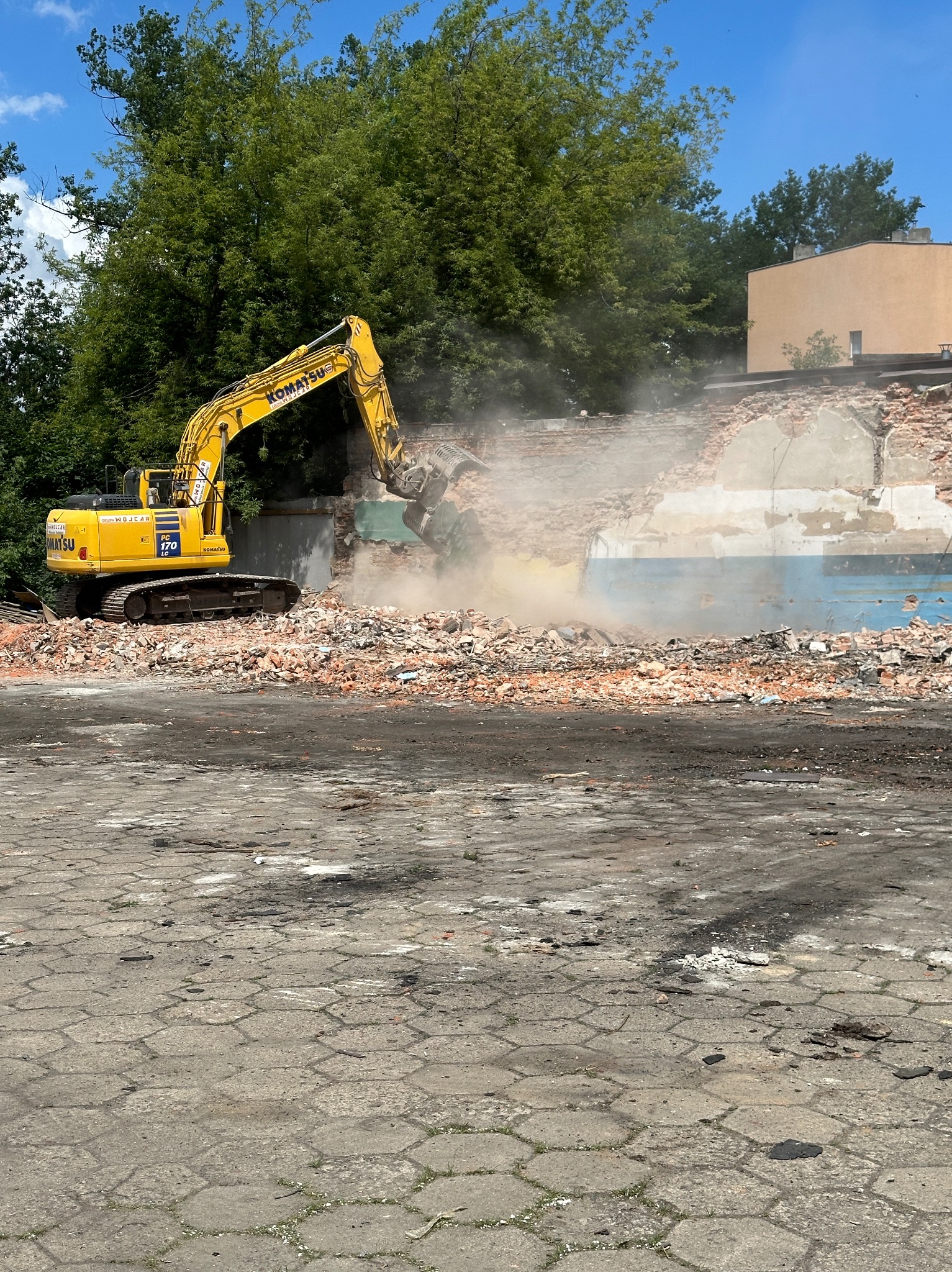 Żółta koparka Komatsu PC 170 LC kruszy resztki muru ceglanego budynku, unosząc tumany kurzu na tle zieleni drzew i błękitnego nieba.