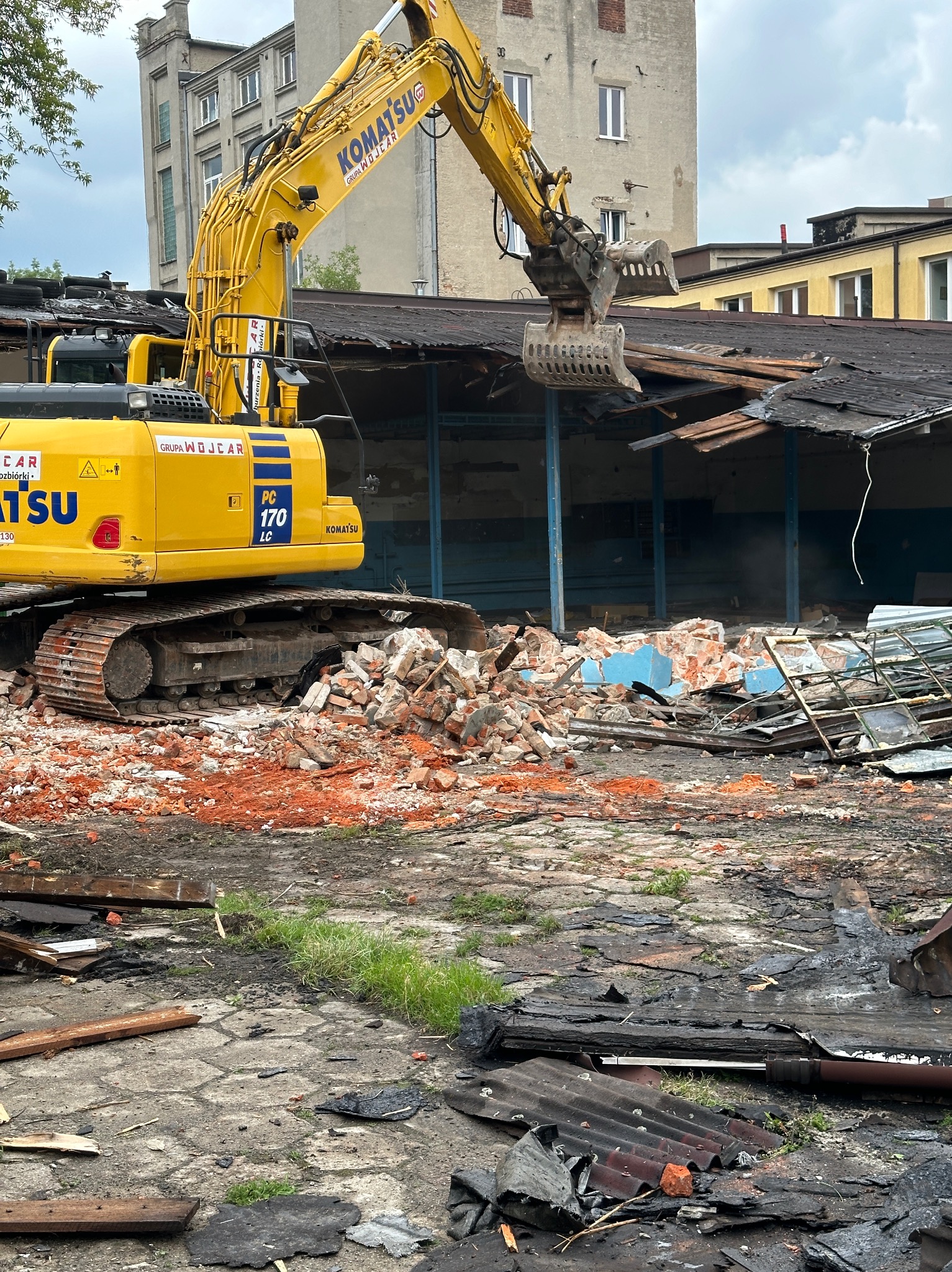 Żółta koparka Komatsu PC170 LC z logo Grupa Wojcar chwyta zdemontowany fragment dachu budynku, w tle ceglany gruz i fragmenty ścian oraz budynki.