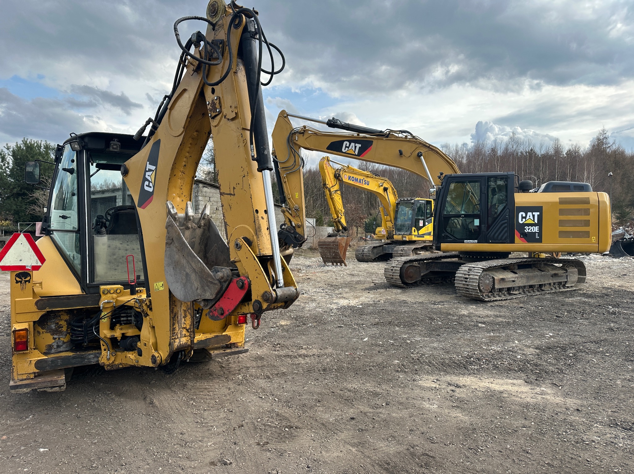 Żółte koparki CAT i Komatsu ustawione na ziemi pod pochmurnym niebem, widoczny tył koparko-ładowarki z naklejką czaszki na znaku ostrzegawczym.