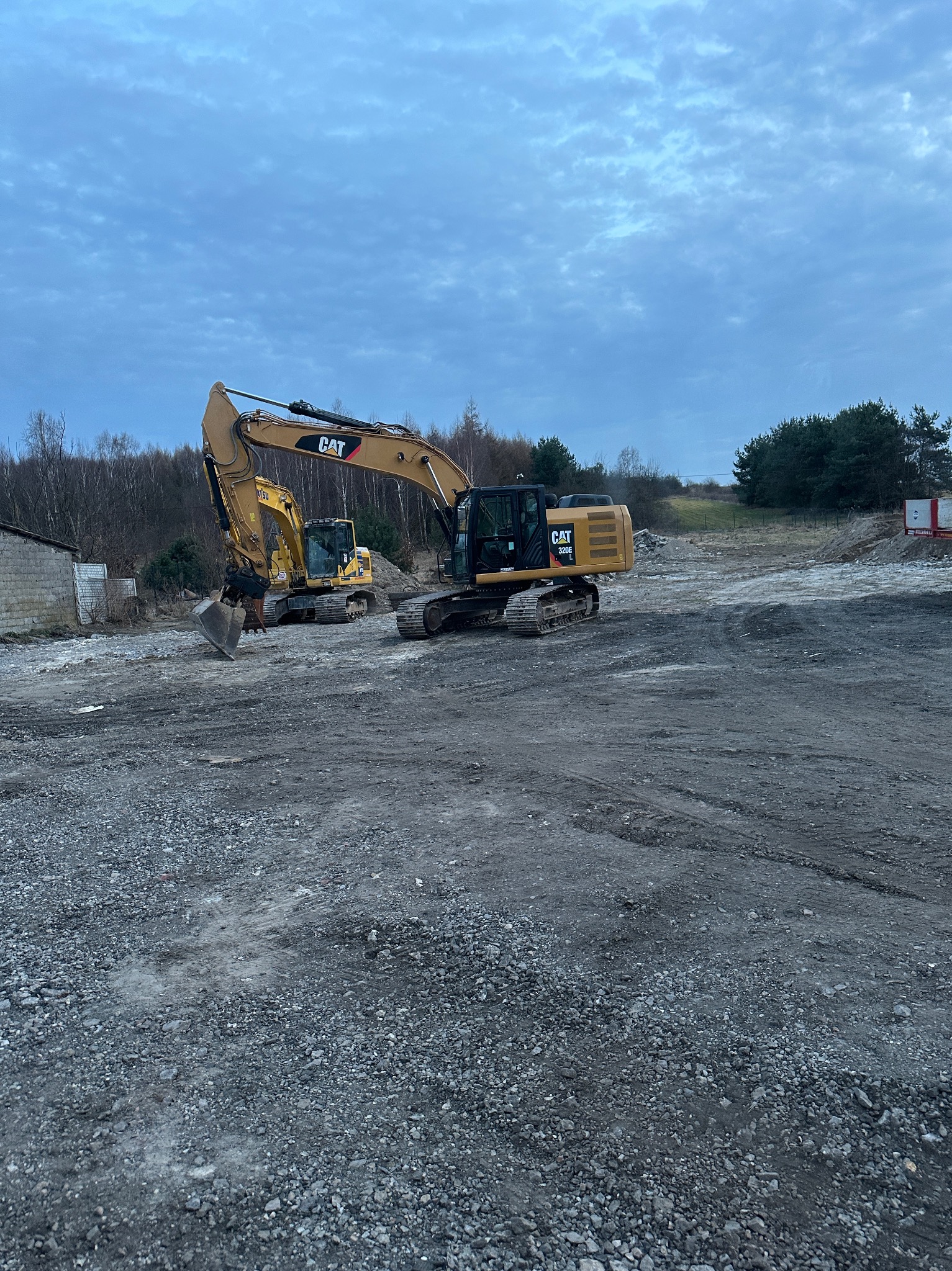 Dwa żółte koparki marki CAT na placu budowy z gruzem i ziemią, pochmurne niebo w tle.