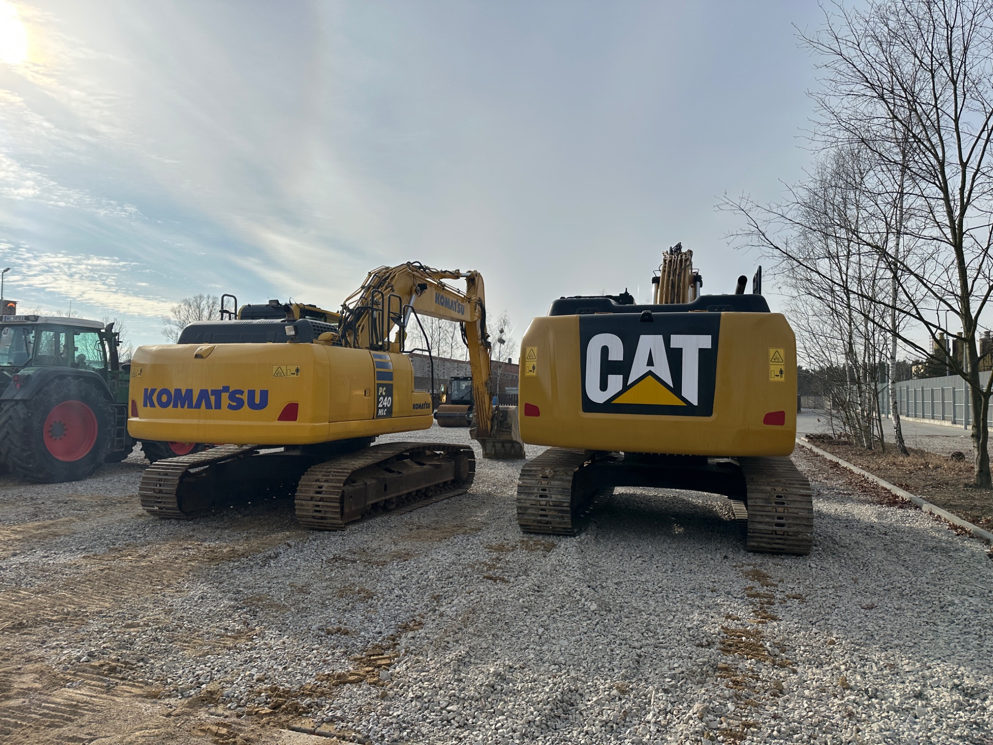 Dwa żółte spychacze: Komatsu PC 240 NLC i Caterpillar, stojące na żwirowym placu w słoneczny dzień, z widocznym ciągnikiem rolniczym z tyłu po lewej stronie.