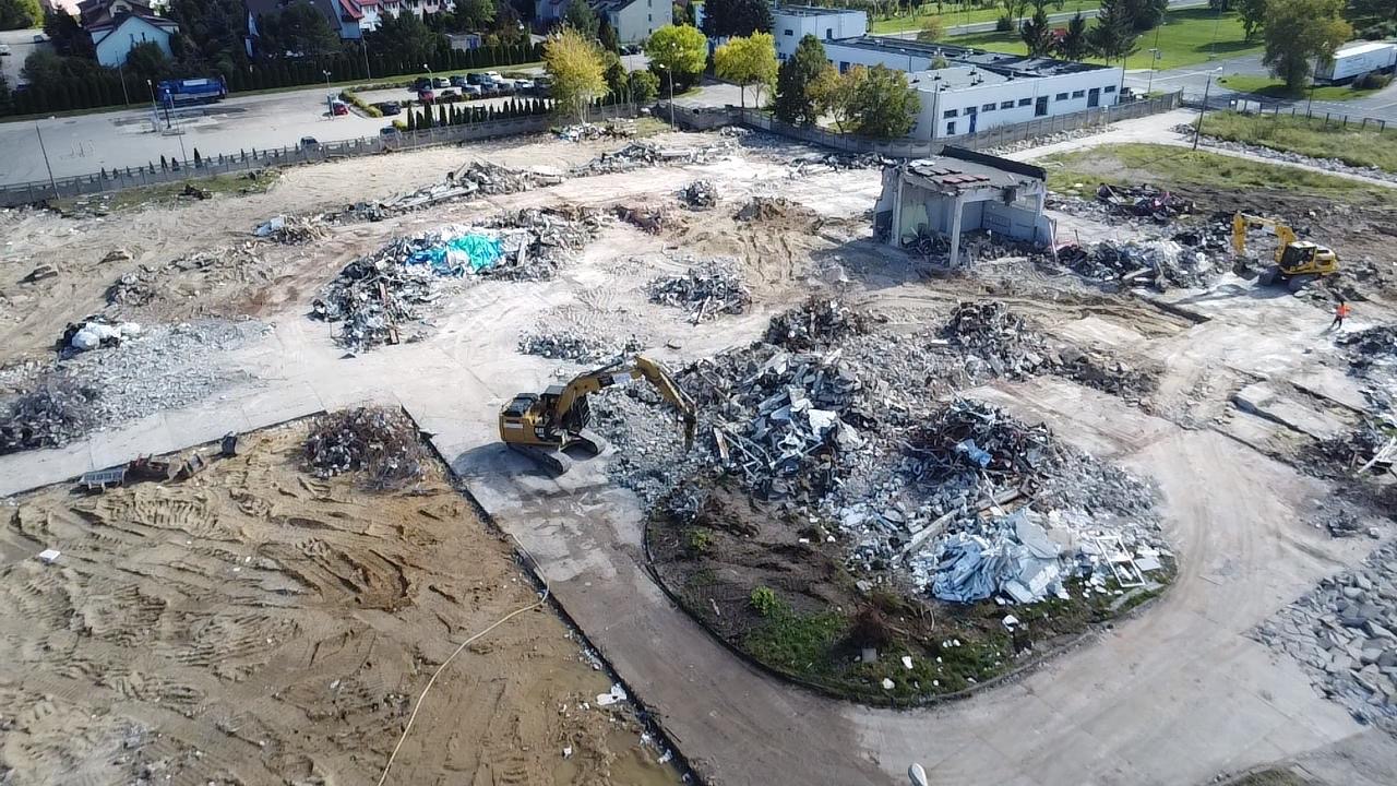 Widok z lotu ptaka na teren po rozbiórce budynku z gruzem, pracującą koparką i fragmentem ocalałej konstrukcji. W tle zabudowa miejska i parking.