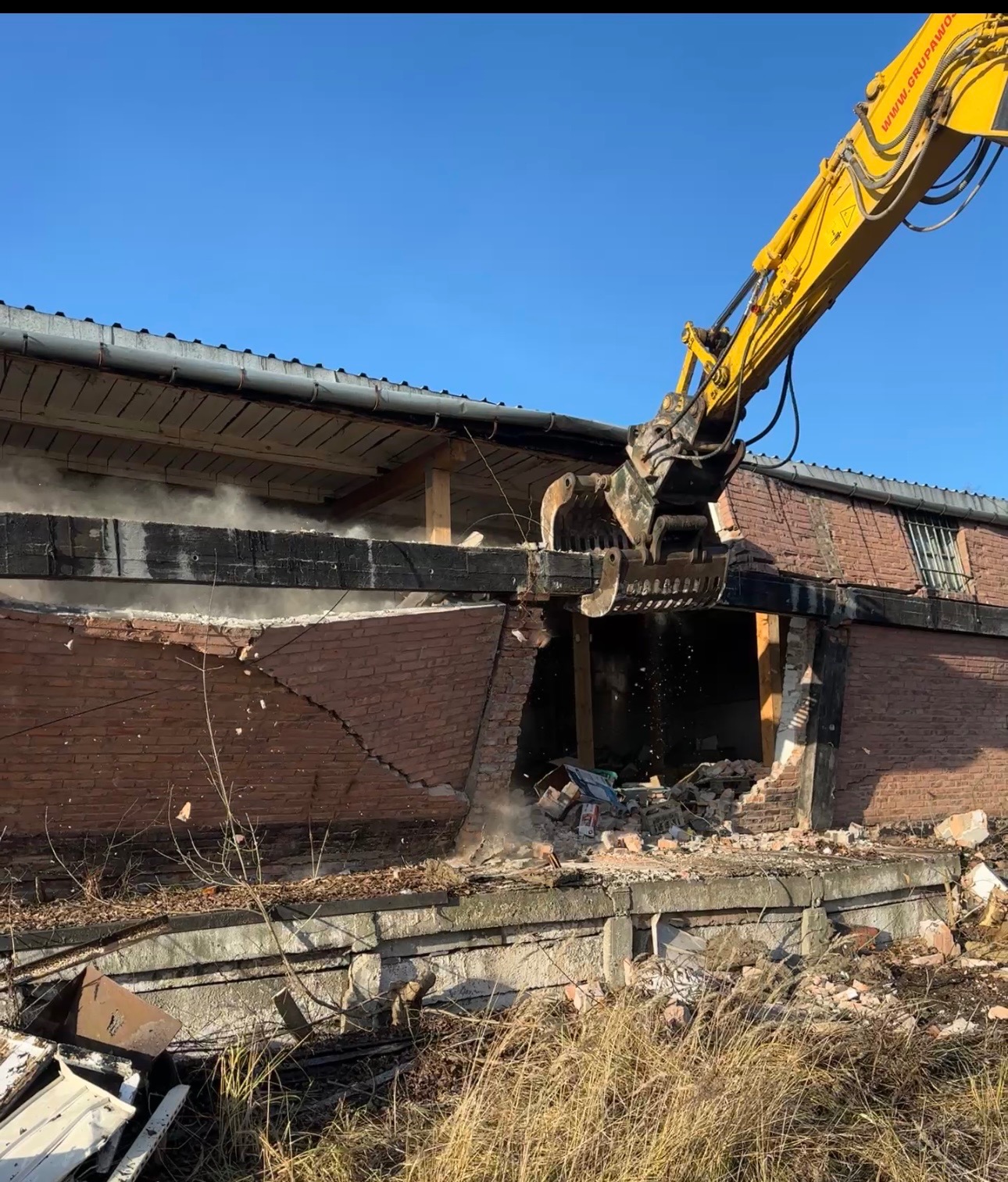 Żółta koparka z precyzją wyburza ceglany budynek, unosząc tumany kurzu. Widoczne zniszczenia i gruz. Częściowo zburzona ściana budynku w słoneczny dzień.