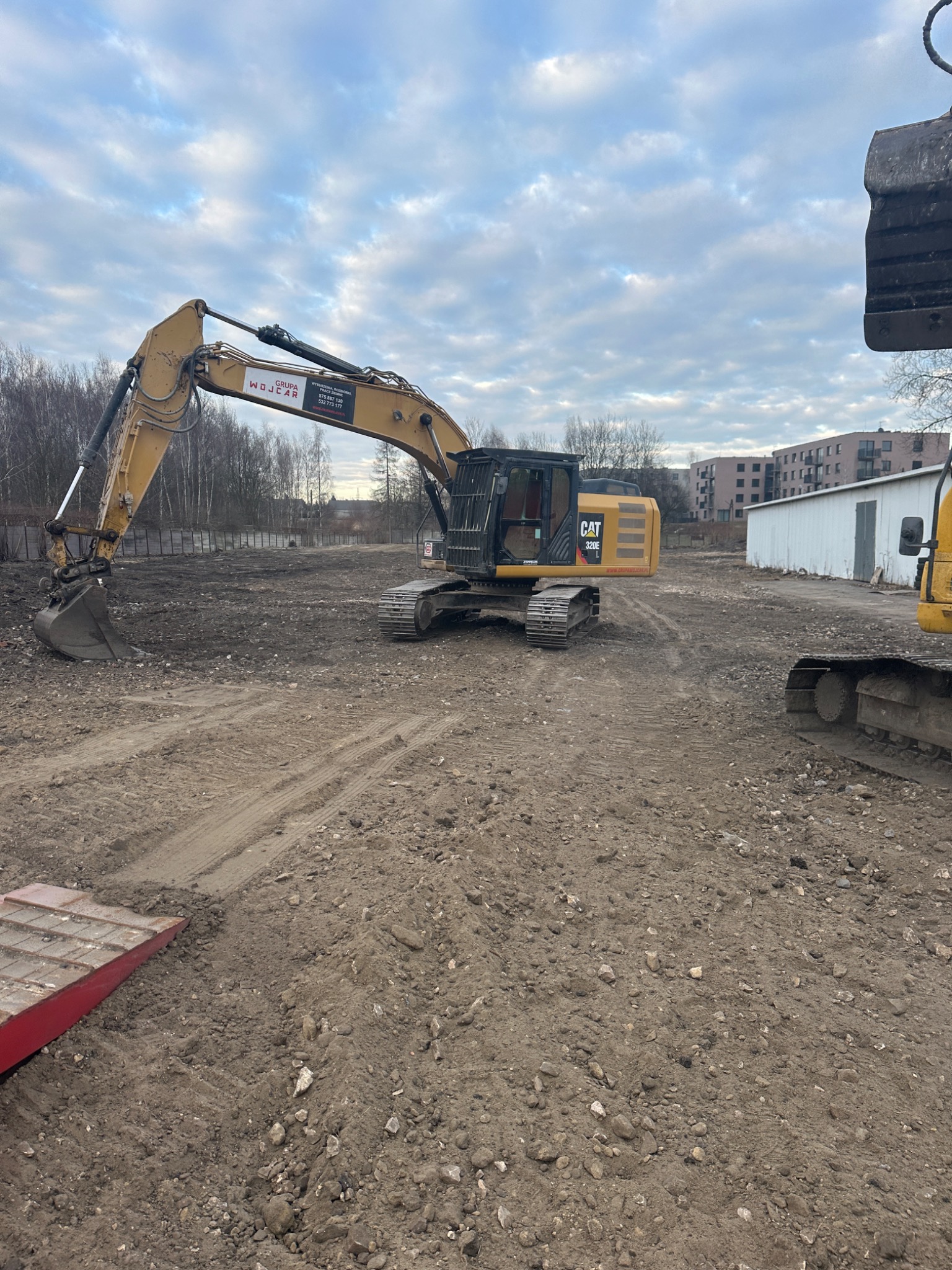 Żółta koparka CAT 320E na placu budowy z budynkami w tle, widoczne ślady opon na ziemi. Na ramieniu koparki naklejka z logo firmy.