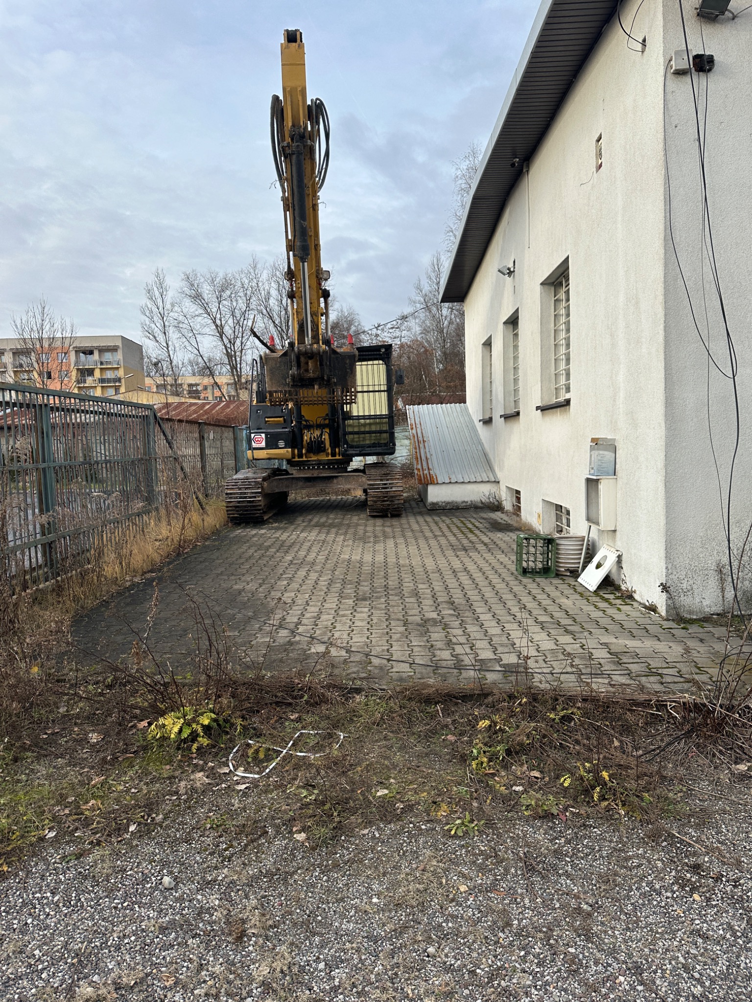 Żółto-czarna koparka na tle białego budynku z oknami i szarej kostki brukowej. Widoczne ogrodzenie i gruz. Pochmurne niebo.