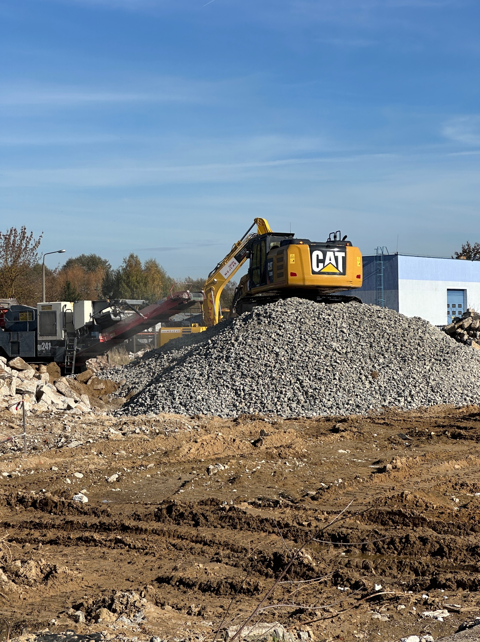 Żółta koparka CAT na szczycie hałdy gruzu, w tle maszyna do kruszenia i budynek. Prace ziemne w toku, błękitne niebo.