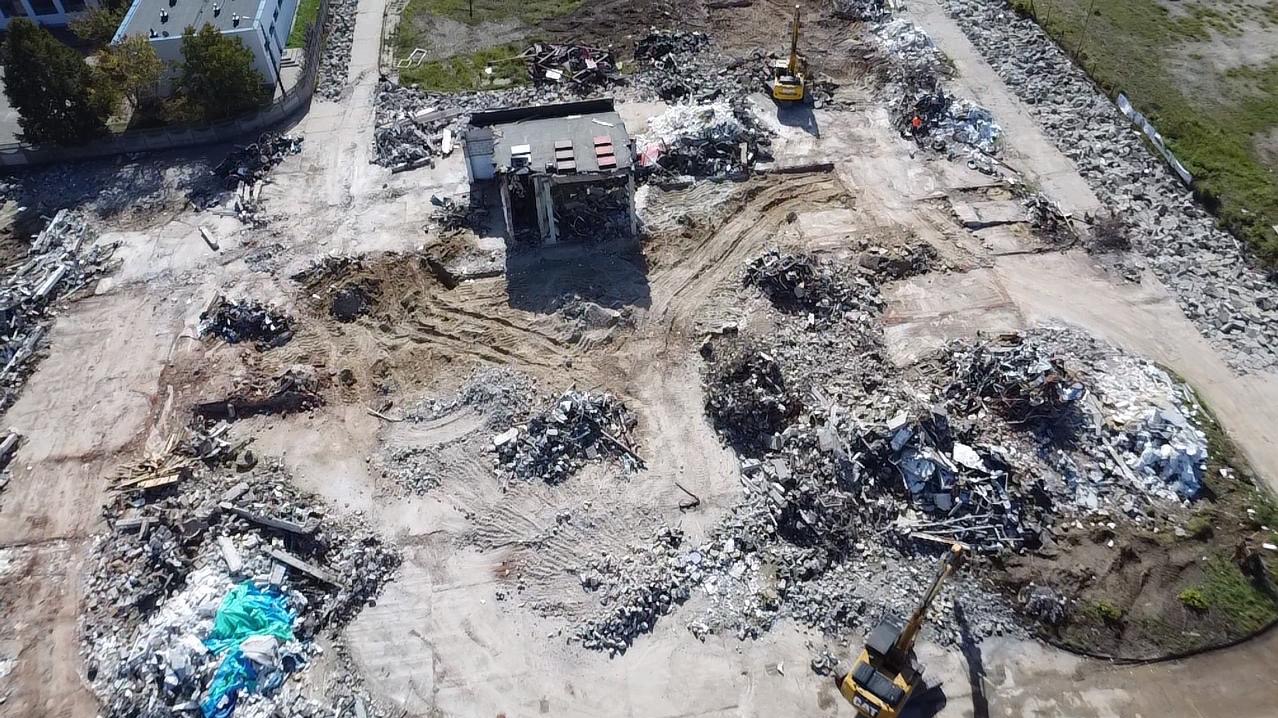 Widok z góry na teren po rozbiórce budynku. Gruz, fragmenty konstrukcji i ciężki sprzęt. Żółta koparka w prawym dolnym rogu. Częściowo zachowane fundamenty.