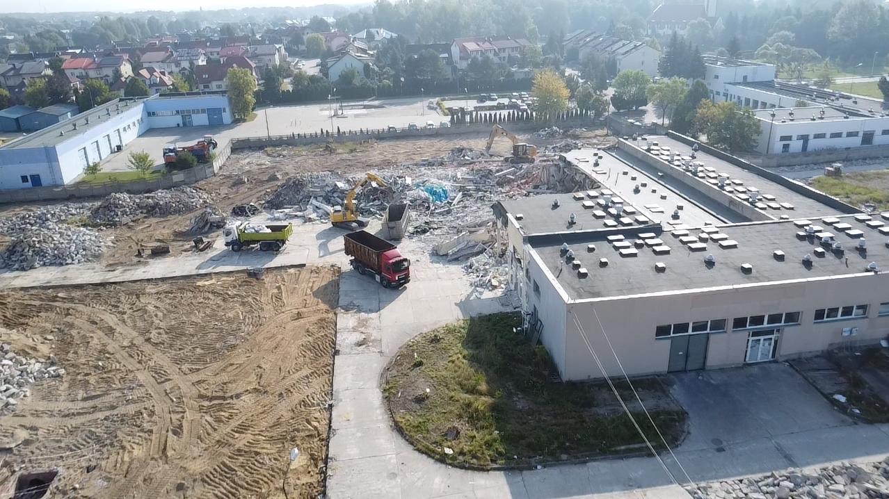 Rozbiórka budynku z użyciem ciężkiego sprzętu: koparki i wywrotki. Widok z góry na plac budowy w trakcie prac rozbiórkowych. Gruz i ziemia.