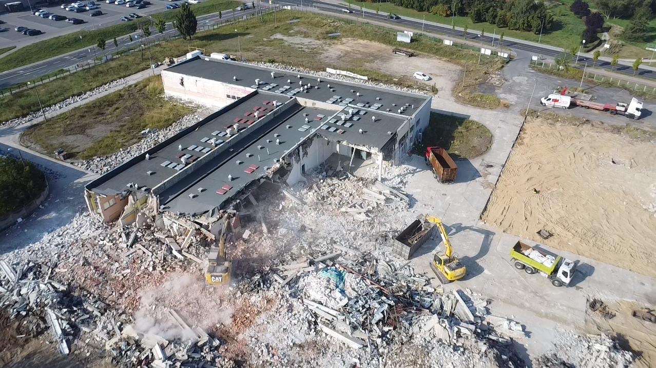 Rozbiórka budynku z lotu ptaka. Widoczne maszyny budowlane i gruzowisko. Częściowo zburzony budynek z odsłoniętymi elementami konstrukcyjnymi.