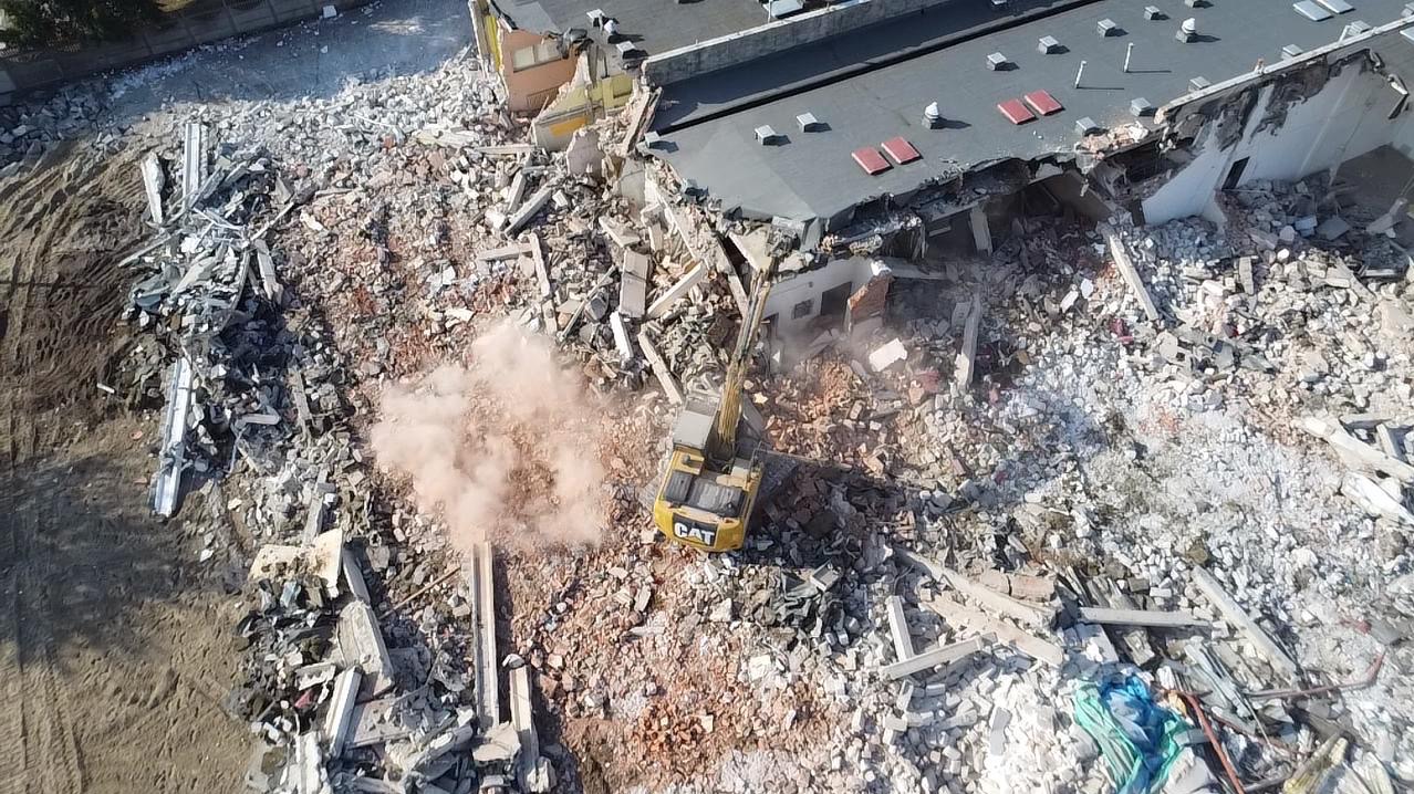 Żółta koparka CAT rozbiera budynek z czerwonym dachem, widok z góry, chmura pyłu. Gruzowisko po rozbiórce.