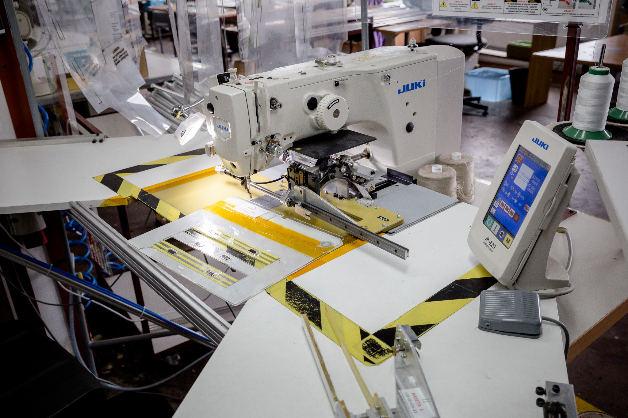 Przemysłowa maszyna szwalnicza JUKI z panelem sterującym, ustawiona na stole z żółto-czarnymi pasami ostrzegawczymi, w tle widoczne szpule nici i wzory wykrojów.