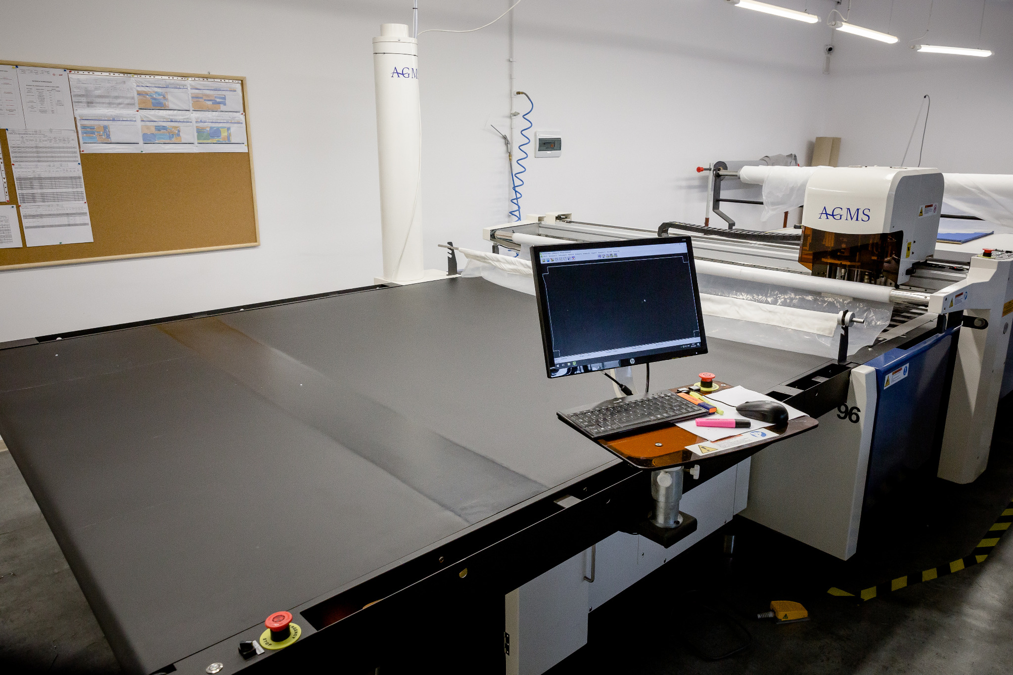 Automatyczna krajalnica tekstylna AGMS z komputerem sterującym, widoczna duża powierzchnia robocza z czarnym materiałem oraz rolka białego materiału na górze, tablica korkowa z dokumentami w tle.
