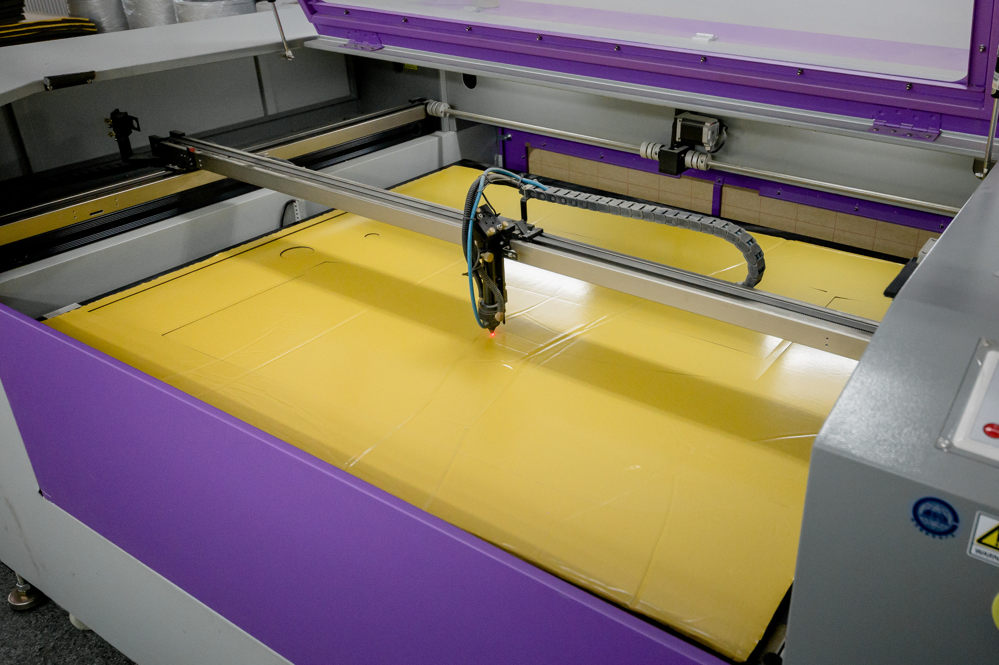 Laserowa maszyna tnąca obrabia arkusz żółtego materiału, widoczny czerwony punkt lasera, maszyna w kolorach fioletowym i szarym.