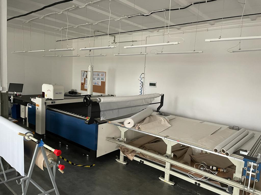 Duże, profesjonalne stanowisko do krojenia materiałów z rolki w jasnym pomieszczeniu, widoczne maszyny i materiały w odcieniach bieli i beżu.