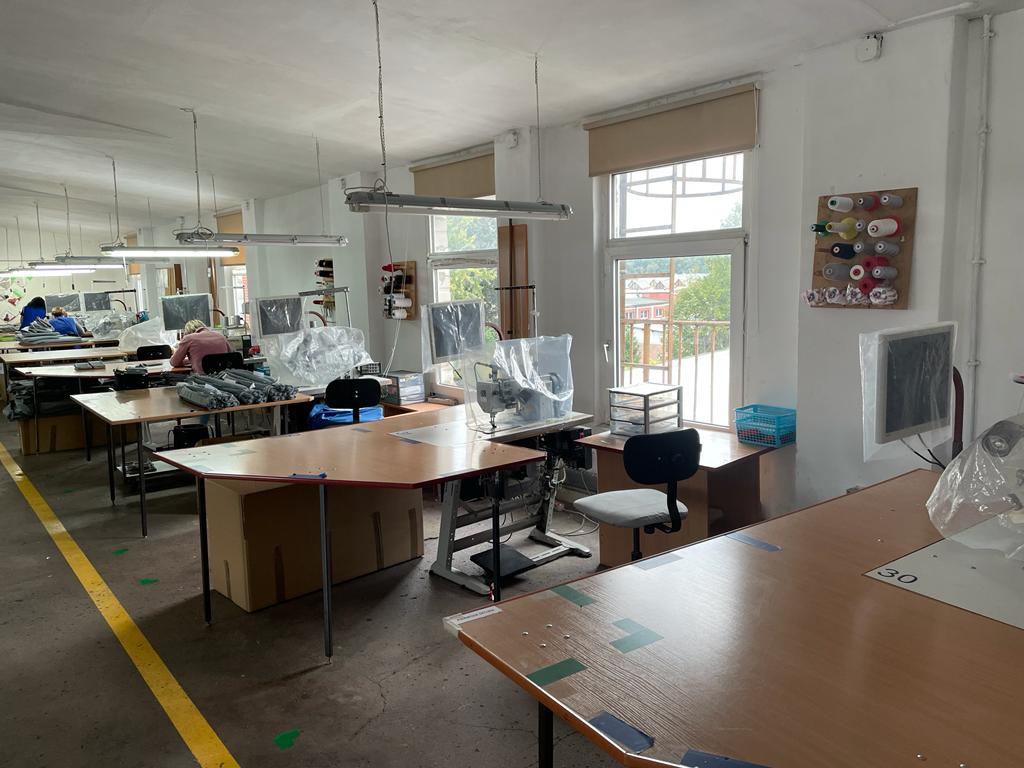 Jasne wnętrze szwalni z rzędami stanowisk pracy, maszynami do szycia przykrytymi folią, widokiem na zieleń za oknem i żółtą linią na betonowej podłodze