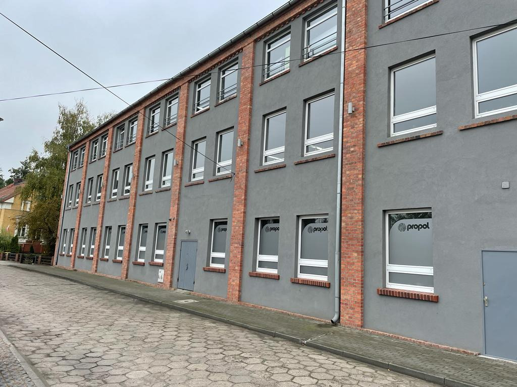 Szary budynek z ceglanymi elementami i oknami z naklejonym logo firmy, widok z ulicy wyłożonej kostką.