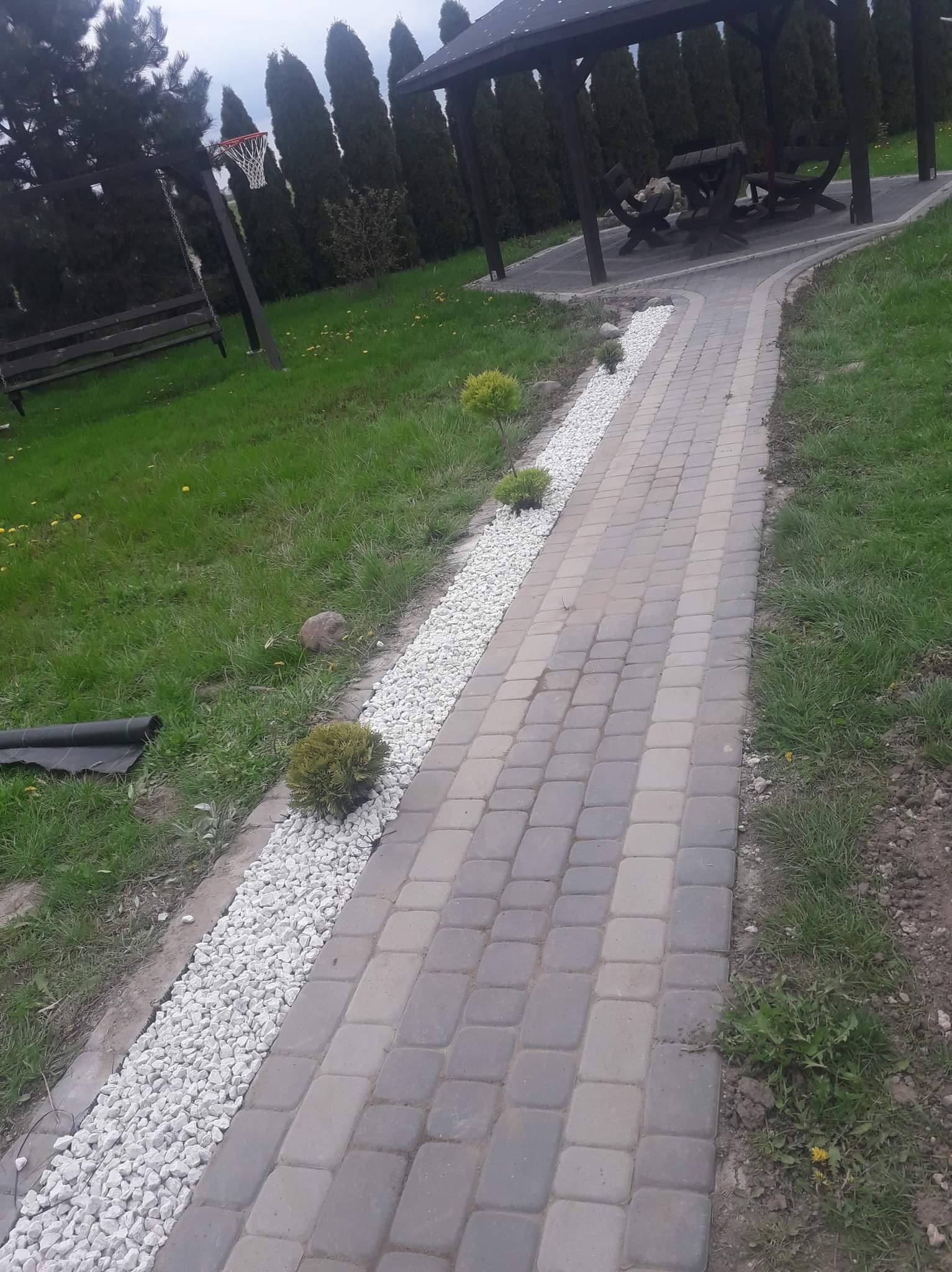 Ścieżka z kostki brukowej z białym kamieniem po bokach, prowadząca do altany z drewnianymi meblami ogrodowymi, w tle wysokie tuje i huśtawka z koszem do koszykówki.