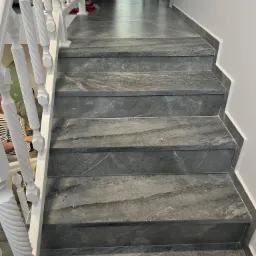 Schody wyłożone szarymi płytkami imitującymi marmur, z białymi, toczonymi balustradami. Widoczny fragment ściany z listwą przypodłogową z płytek.