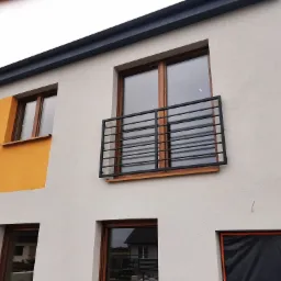 Czarna, metalowa balustrada balkonowa z poziomych prętów, zamontowana na nowoczesnym budynku z oknami w drewnianych ramach i fragmentami pomalowanymi na żółto.