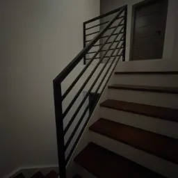 Nowoczesne schody drewniane z czarną, minimalistyczną balustradą poziomą, oświetlone wpadającym światłem z góry, z widocznymi drzwiami w tle.