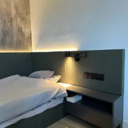 Nowoczesne łóżko z tapicerowanym zagłówkiem w odcieniach szarości, z wbudowaną szafką nocną i oświetleniem, na tle ściany z tapetą w motywie leśnym.