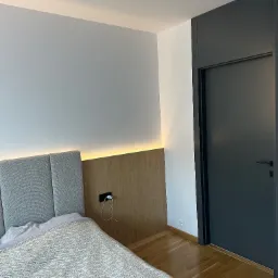 Nowoczesna sypialnia z szarą tapicerowaną ścianą za łóżkiem, drewnianą boazerią z oświetleniem LED, parkietem i ciemnymi drzwiami w zabudowie.