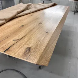 Blat drewniany z wyraźnym usłojeniem, częściowo przykryty papierem ochronnym w warsztacie stolarskim, widoczne niedoskonałości drewna.