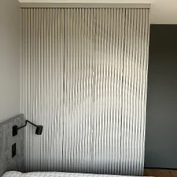 Szara szafa przesuwna z lamelami w sypialni, widoczna zza wezgłowia łóżka z pikowaną narzutą, obok ściany z lampką.