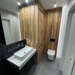 Nowoczesna łazienka w Nowym Opolu z drewnianą zabudową, umywalką nablatową na czarno-białym postumencie i podświetleniem LED, widok z perspektywy kabiny prysznicowej.