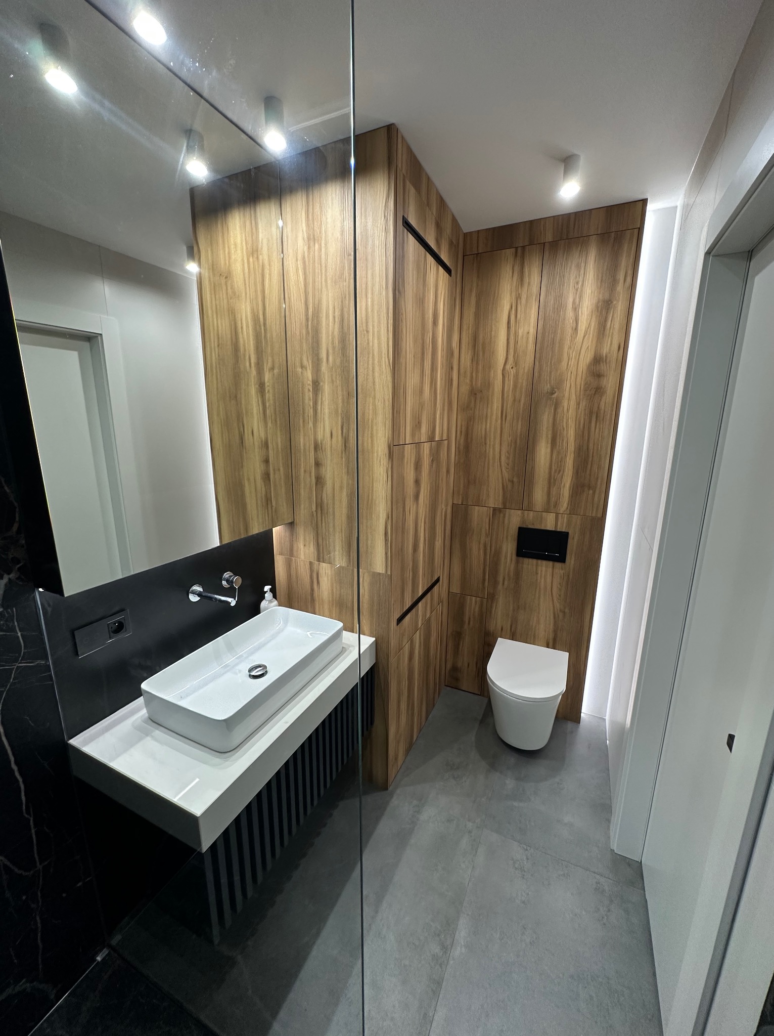 Nowoczesna łazienka w Nowym Opolu z drewnianą zabudową, umywalką nablatową na czarno-białym postumencie i podświetleniem LED, widok z perspektywy kabiny prysznicowej.