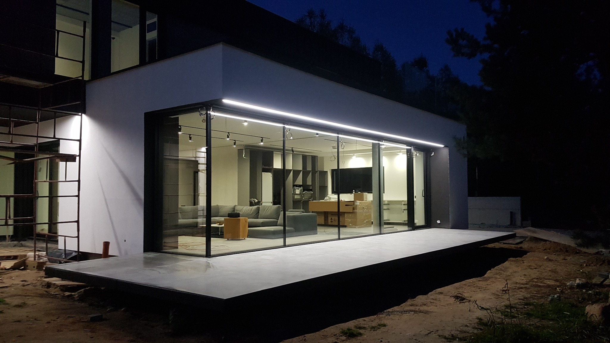 Nowoczesny dom z dużymi przesuwnymi oknami, widok na salon z szarą sofą, elementami drewnianymi i kartonami, teren wokół domu w trakcie prac wykończeniowych, widoczne rusztowanie.