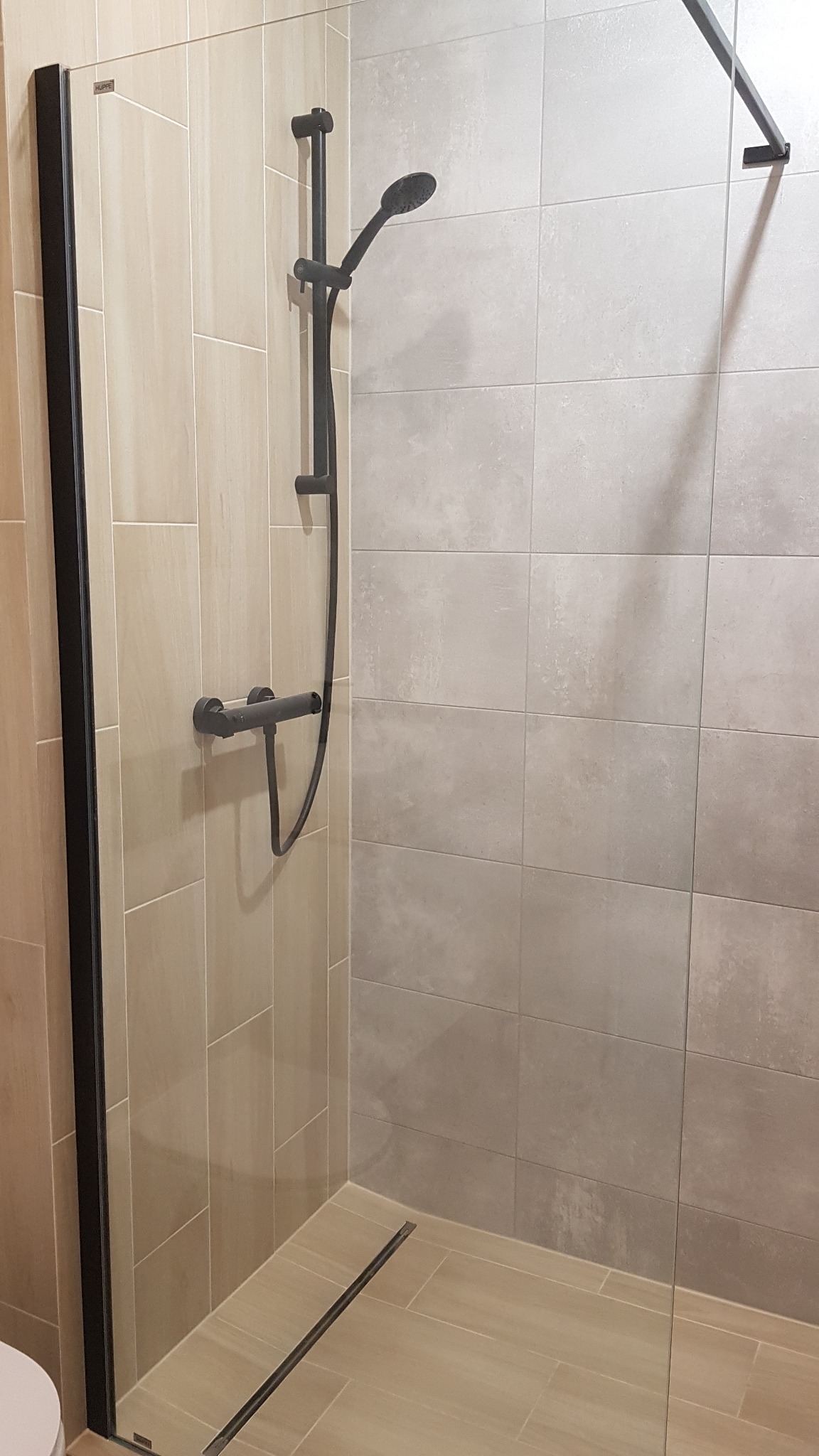 Nowoczesna kabina prysznicowa z czarną armaturą, przeszklona, z odpływem liniowym w podłodze wyłożonej beżowymi płytkami imitującymi drewno, ściany wykończone płytkami w dwóch odcieniach szarości.