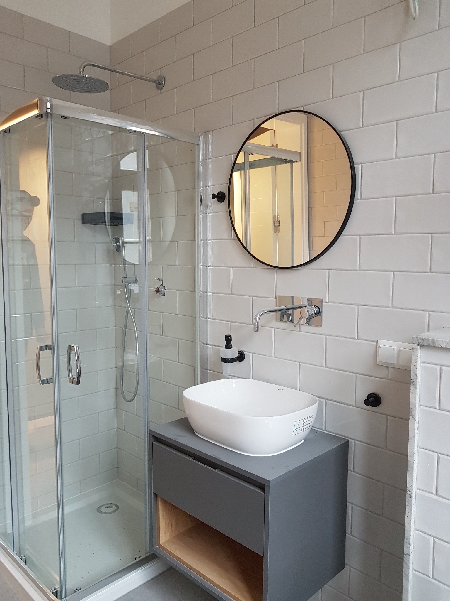 Nowoczesna łazienka z kabiną prysznicową, okrągłym lustrem i umywalką nablatową na szarej szafce z drewnianą półką, wykończona białymi, prostokątnymi płytkami.