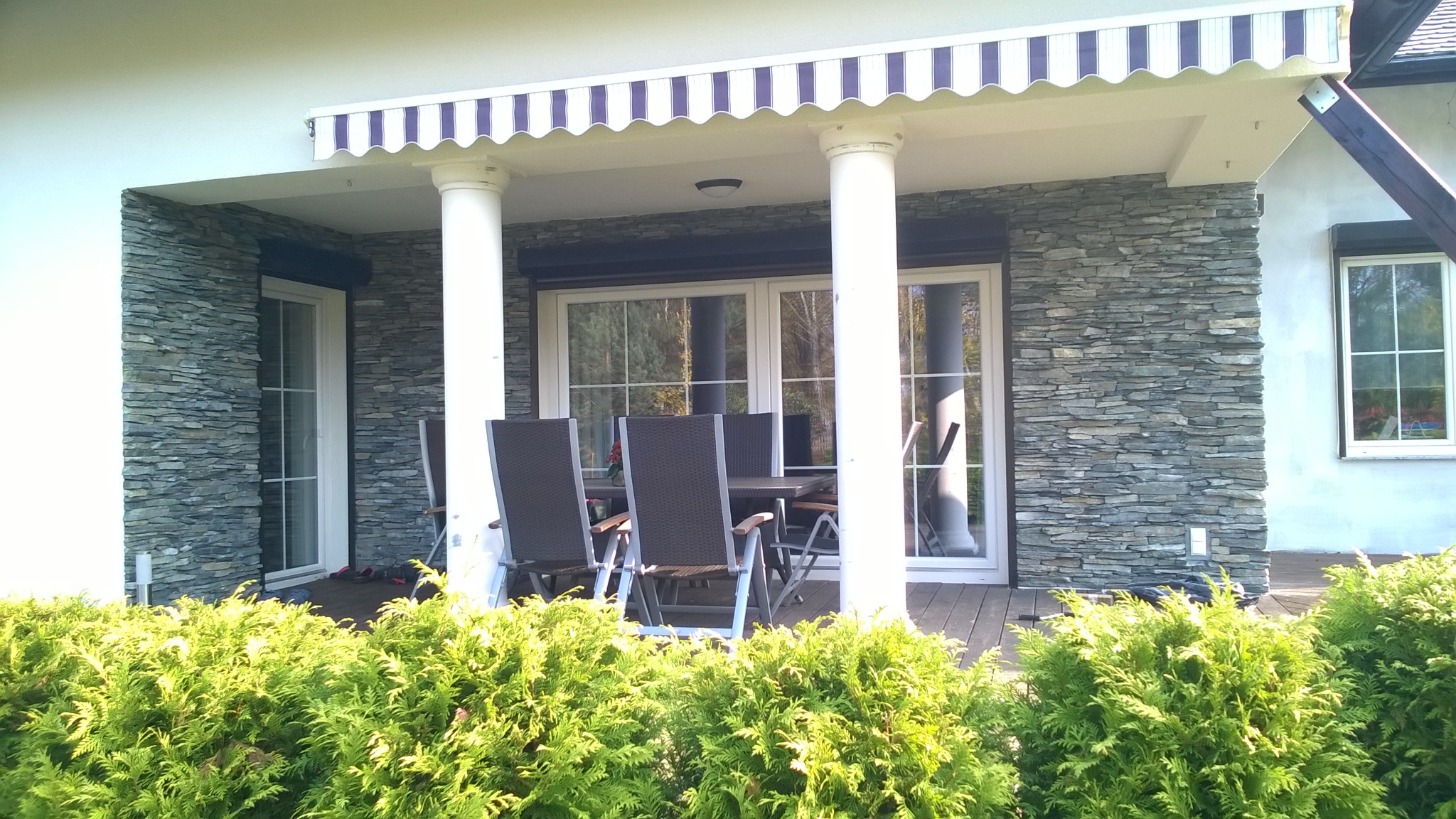 Zadaszony taras z kamienną elewacją, białymi kolumnami i markizą w biało-niebieskie paski, widok zza zielonych krzewów.