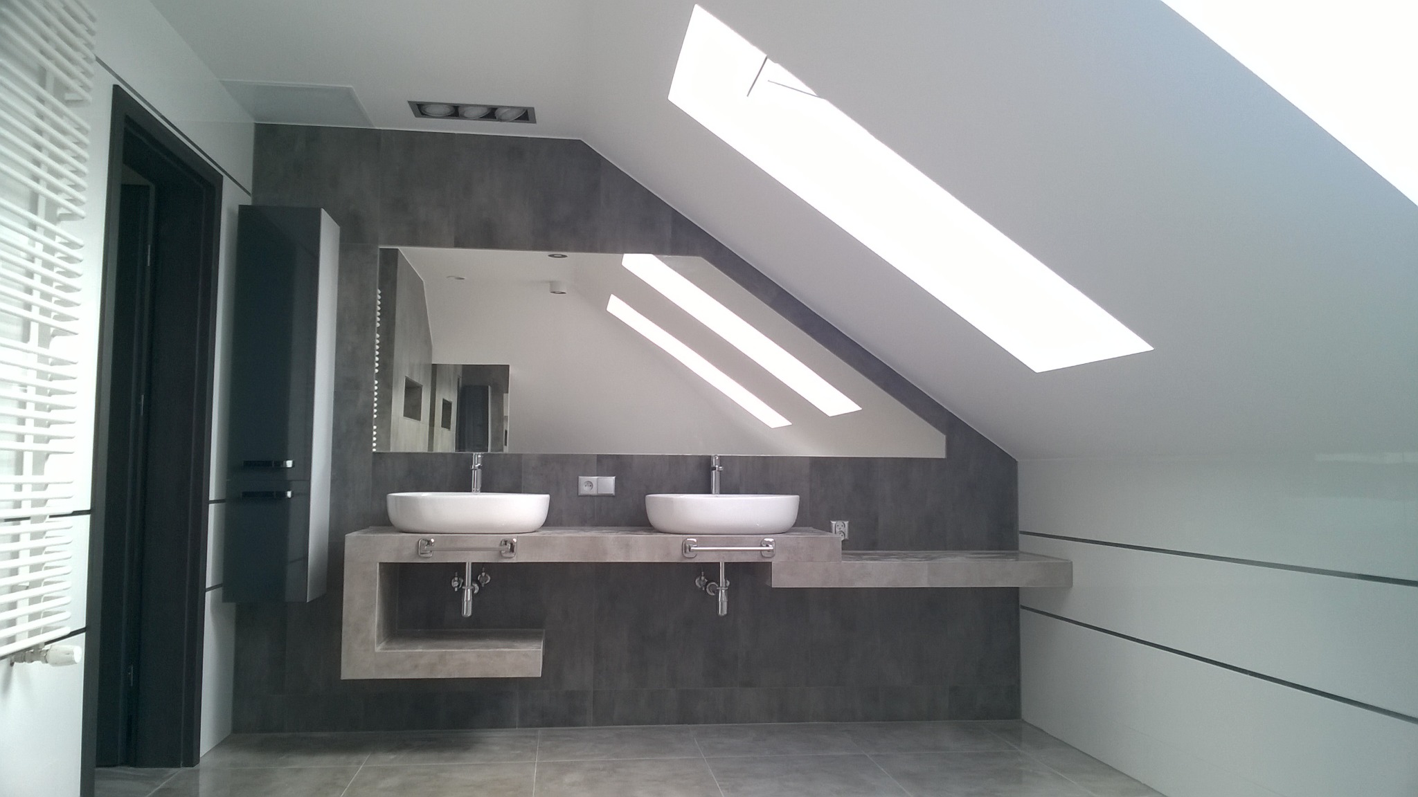 Nowoczesna łazienka na poddaszu z dwoma umywalkami nablatowymi, szarą zabudową i dużym lustrem, oświetlona przez okno dachowe.