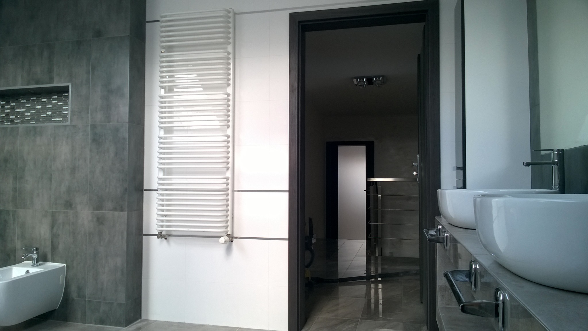 Nowoczesna łazienka z szarymi płytkami, białym grzejnikiem drabinkowym, wiszącym bidetem i umywalkami nablatowymi, widok z otwartych drzwi na dalszą część pomieszczenia.