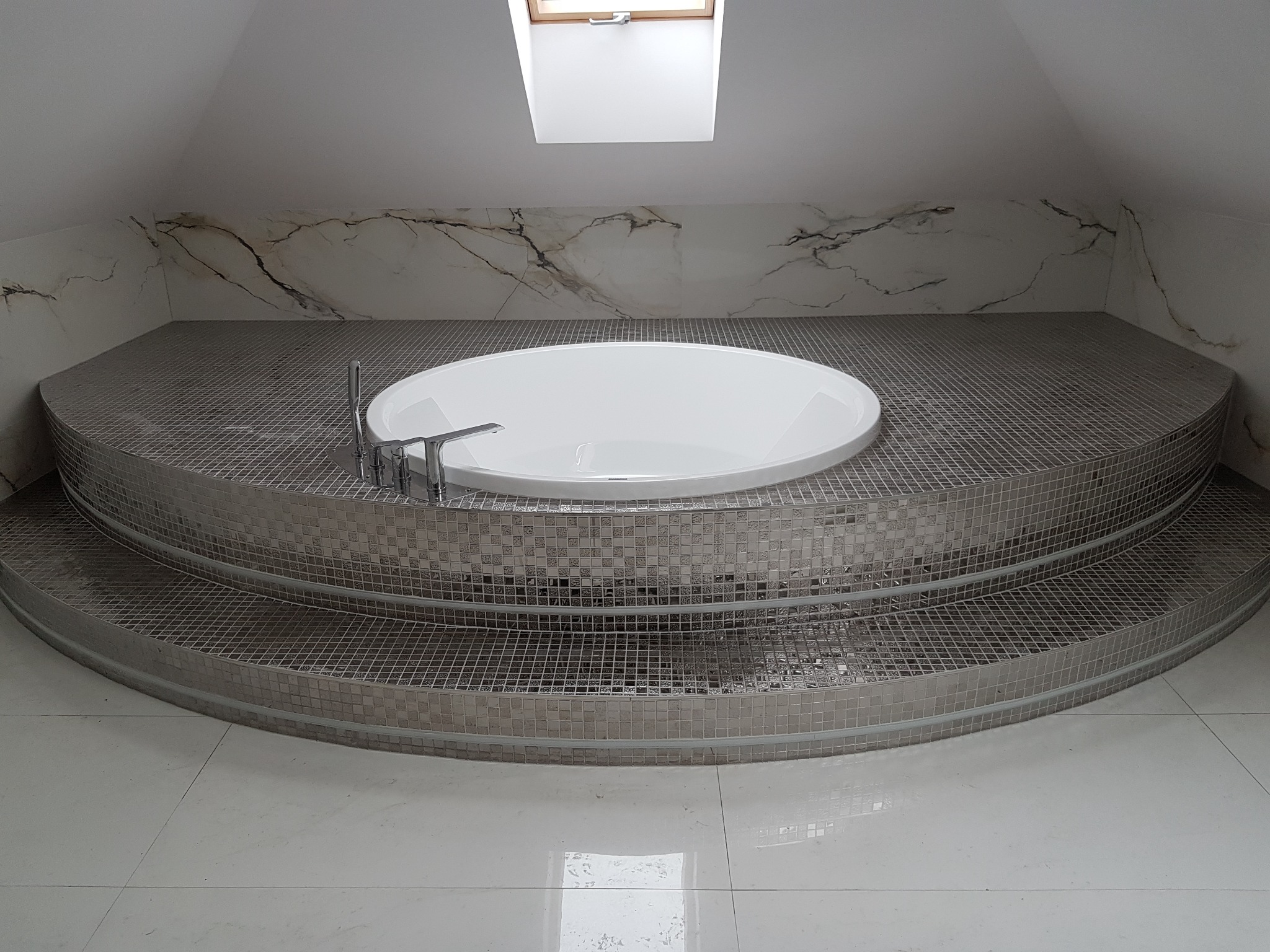 Nowoczesna, owalna wanna na podwyższeniu wykończonym mozaiką w łazience na poddaszu z oknem dachowym.
