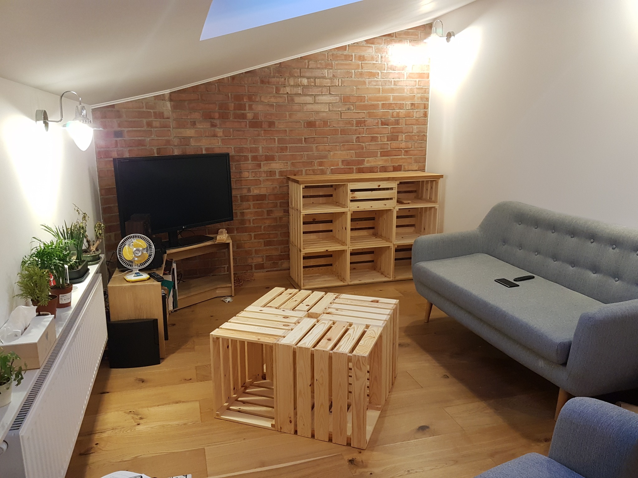 Jasne wnętrze pokoju na poddaszu z ceglaną ścianą, drewnianą podłogą i meblami z surowego drewna: regał, stolik kawowy z drewnianych skrzynek, szara sofa z pilotami, grzejnik z roślinami.