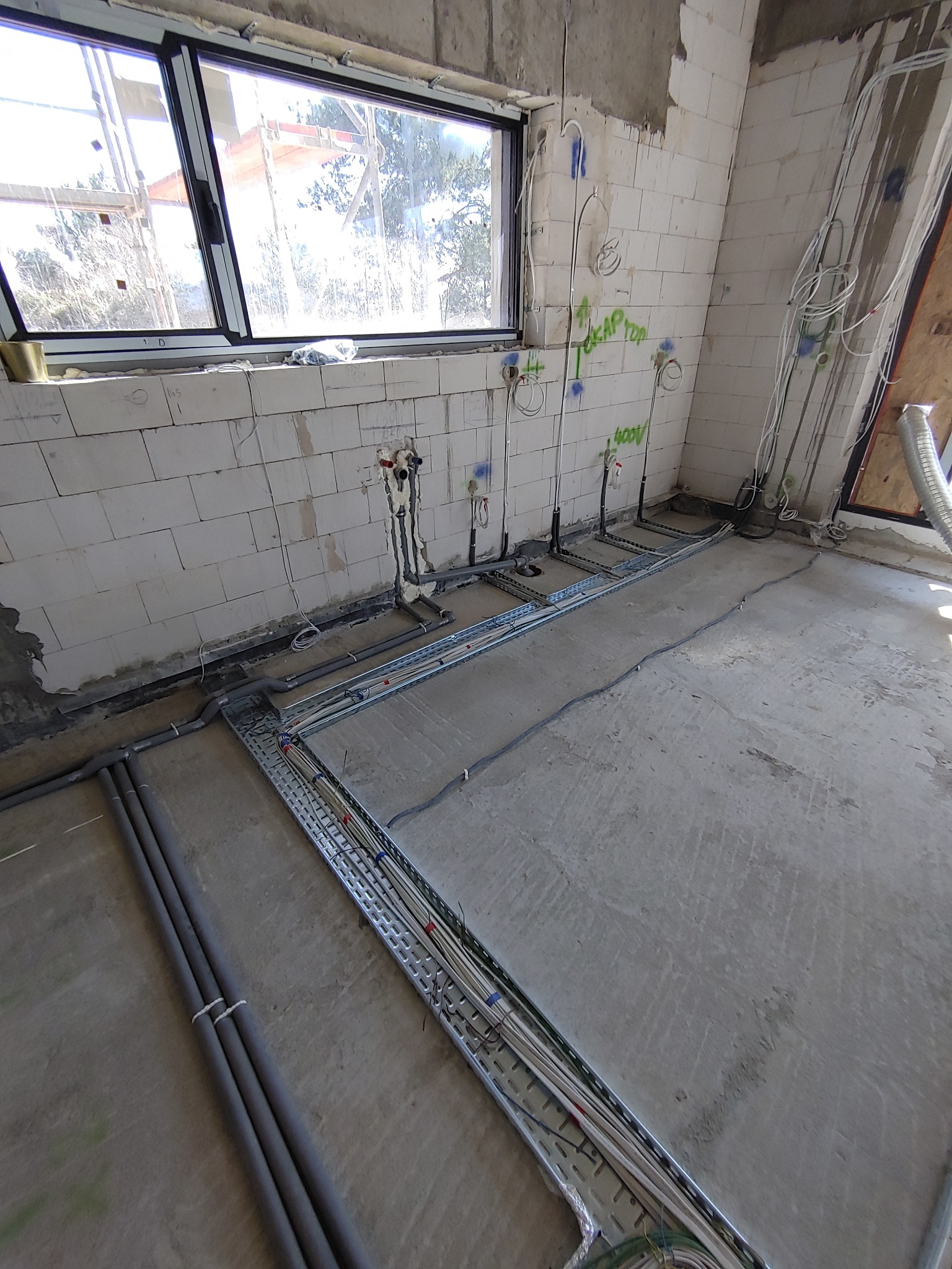 Surowy stan wnętrza w trakcie prac instalacyjnych: widoczne kable, rury, puszki elektryczne i instalacje wodno-kanalizacyjne na ścianach z betonu komórkowego oraz koryta kablowe na podłodze.