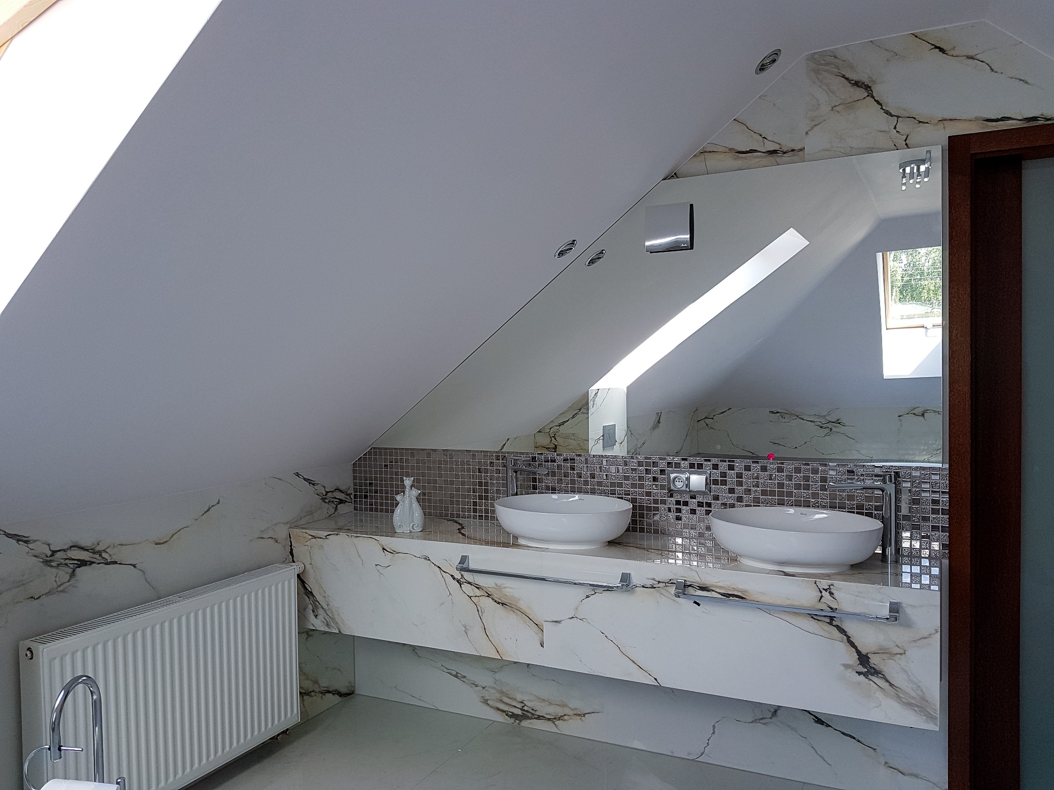 Nowoczesna łazienka na poddaszu ze skośnym sufitem, marmurowa umywalka nablatowa z dwoma białymi misami, mozaika na ścianie i grzejnik żeberkowy.