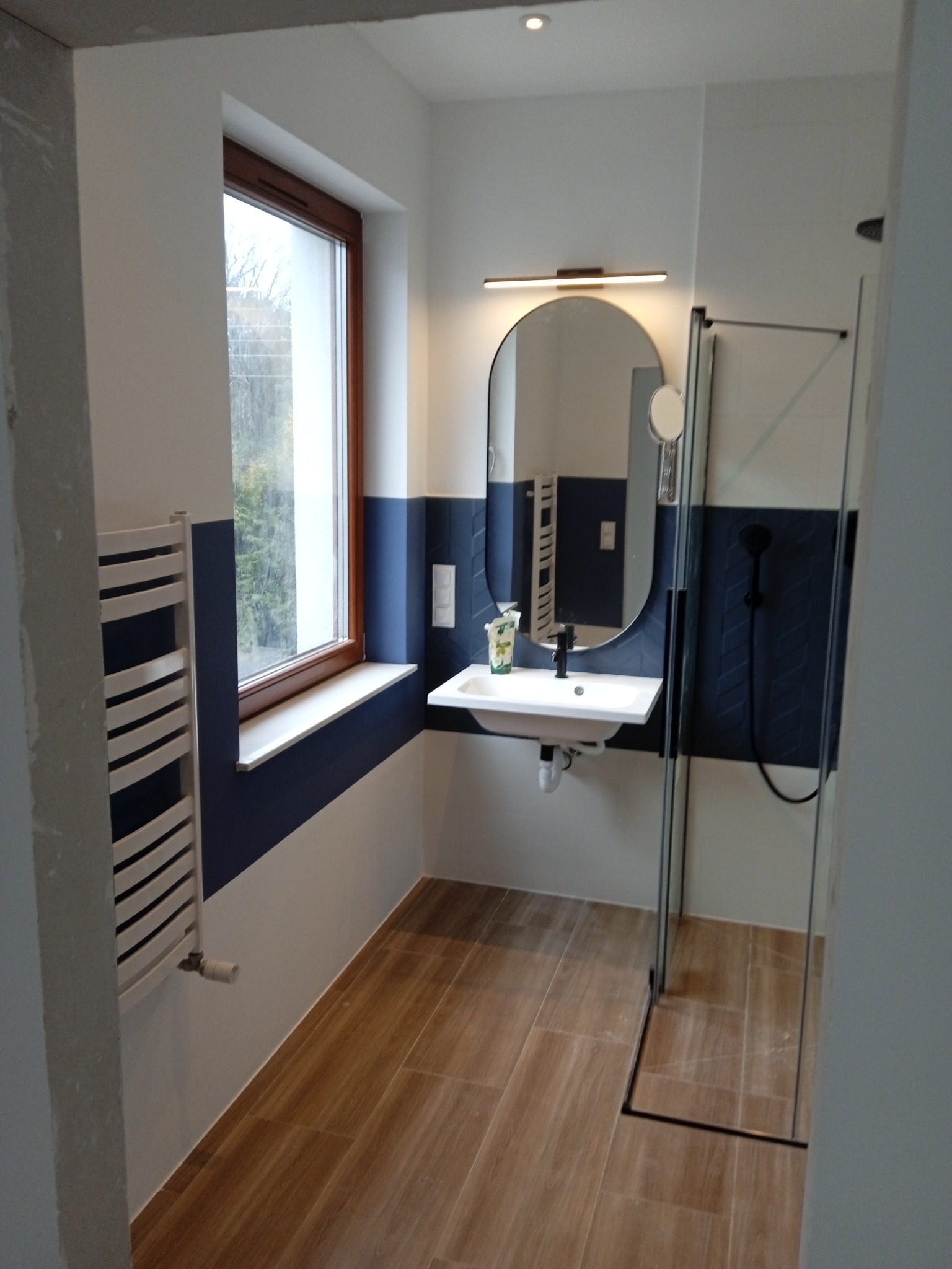 Nowoczesna łazienka z drewnopodobną podłogą, umywalką nablatową z owalnym lustrem podświetlanym listwą LED, kabiną prysznicową ze szklanymi drzwiami i grzejnikiem drabinkowym przy oknie.