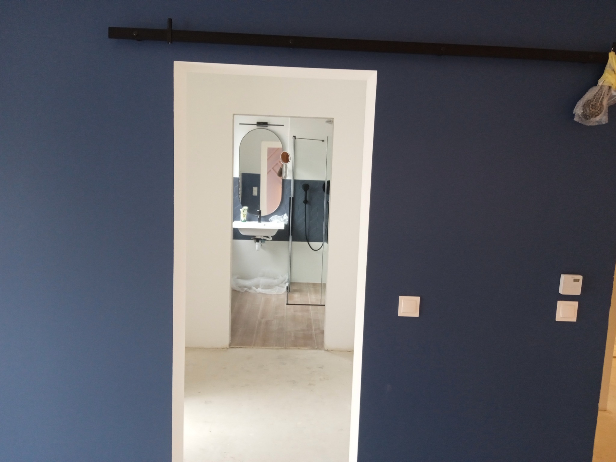 Niebieska ściana z białym otworem drzwiowym prowadzącym do wykończonej łazienki z owalnym lustrem, białą umywalką i kabiną prysznicową; widoczna szyna do drzwi przesuwnych.